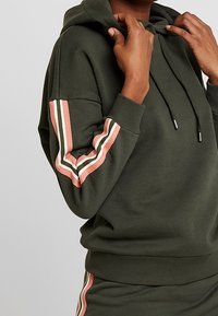 Olijfgroene sweatshirt met capuchon, gestreepte accenten in het roze, wit en groen op de mouwen, en een geribbelde onderrand en manchetten.