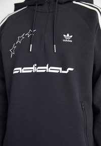 Black Adidas hoodie featuring white logo and star design, drawstring hood, and zippered side pocket with contrasting white stripes.