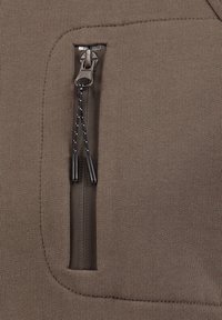 Un bolsillo marrón con cremallera sobre un tejido texturizado, que presenta una cremallera suave con tiradores de cordón negros en contraste y un diseño aerodinámico.