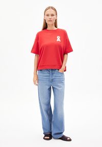 Roter Kurzarm-Sweatshirt mit weißem Band-Logo, kombiniert mit hellblauen, locker sitzenden Jeans und schwarzen Sandalen mit offenen Zehen. Flache, glatte Stoffstruktur.