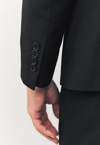 Ærmet på en sort jakkesætjakke med en ren tekstur, fire sorte knapper langs manchetterne og en let buet kant forneden.