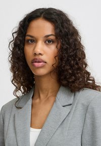 Šedý blazer s výraznými klopy, strukturovanou tkaninou a slim fit střihem, nošený přes bílý žebrovaný top se subtilním designem límce.