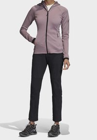 Sudadera morada con cremallera y un diseño ajustado, detalles en negro y capucha. Combinada con pantalones negros y zapatos de senderismo multicolores.