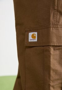 Braune Cargo-Hosen aus strukturiertem Material, mit einer Seitentasche und einem weißen Etikett mit gelbem Logo.