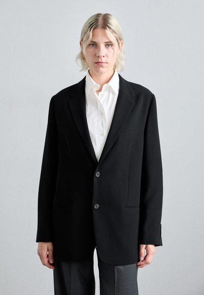 Black blazer with two buttons, made of smooth fabric. Features notched lapels and side pockets, paired with a white shirt and gray trousers.