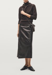 Maglione marrone lavorato a maglia con colletto alto, abbinato a una lunga gonna di pelle nera. Scarpe basse senza lacci completano il look, insieme a una piccola borsa.