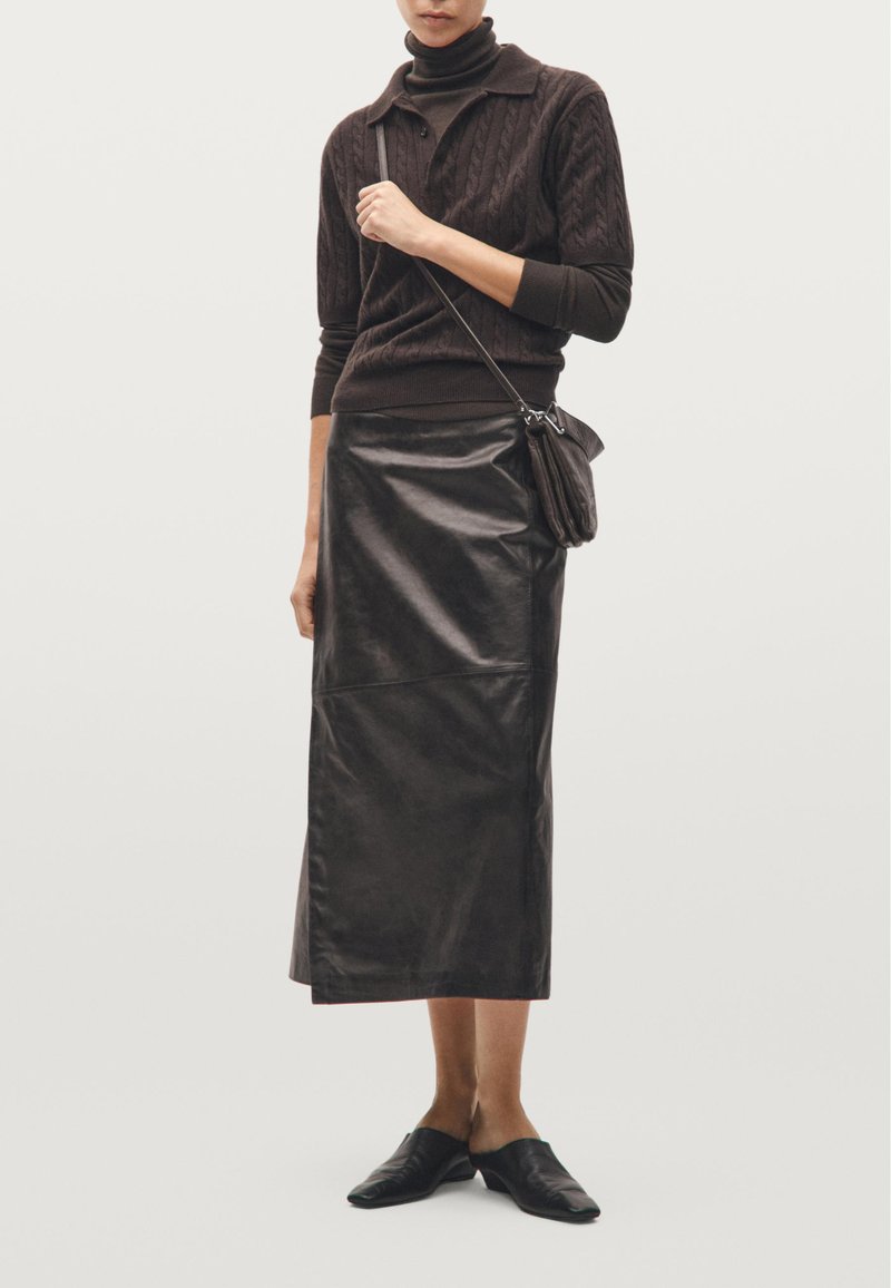Hnědý pletený svetr s vysokým límcem, kombinovaný s dlouhou tmavou koženou sukní. Celkový vzhled doplňují černé ploché slip-on boty a malá taška.