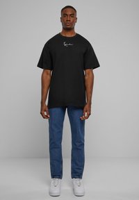 Svart oversized t-shirt med en vit signaturlogga på bröstet, tillsammans med blå denimjeans och vita sneakers på en plain bakgrund.