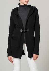 Czarny dzianinowy kardigan z kapturem, z zapięciem z przodu, dwiema kieszeniami i ściągaczami przy mankietach. Noszony na ciemnym golfie i białych spodniach.