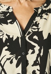 Blouse à motifs floraux en noir et blanc avec un col en V, fermeture à boutons, et une texture de tissu douce ; dotée de manches larges et d'une coupe décontractée.