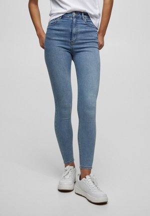 Personne portant un jean skinny taille haute bleu clair et des baskets blanches, debout devant un fond uni.