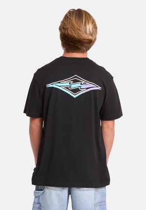 Persona con i capelli castano chiaro che indossa una t-shirt nera con un logo onda blu e viola sulla schiena e jeans azzurri.