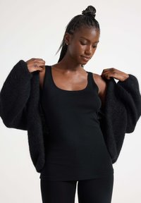 Zwarte oversized cardigan van textuurstof, open gedragen over een aansluitend zwart tanktop. Glad, lichtgewicht materiaal, casual ontwerp en comfortabele pasvorm.