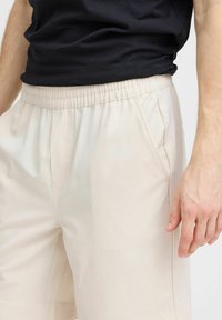 Pantalones beiges con cintura elástica, bolsillos laterales, textura suave y un corte relajado, combinados con una camiseta negra.