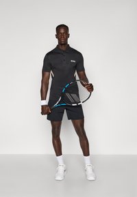 Polo noir côtelé avec logo "BOSS" blanc, associé à un short marine. Le modèle tient une raquette de tennis avec un accent bleu, portant des baskets et des chaussettes blanches.