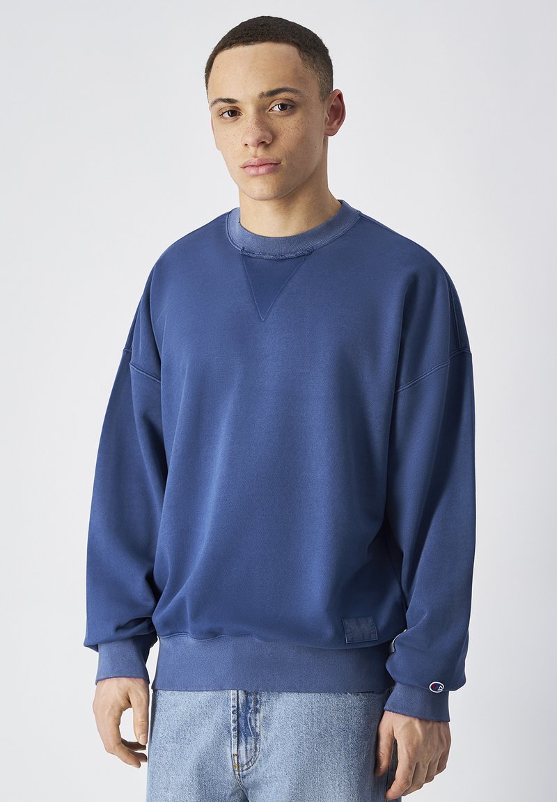 Felpa blu con colletto rotondo, vestibilità oversize, polsini e orlo a coste, texture liscia, con una patch del logo nella parte inferiore sinistra.