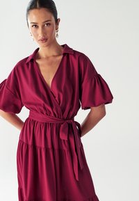 Bordeauxfarbendes Wickelkleid mit kurzen, puffärmeligen Ärmeln, tiefem V-Ausschnitt und gebundenem Taillenband. Verfügt über einen gestuften Rock und einen weichen, strukturierten Stoff.