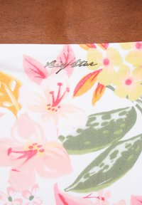 Tkanina z nadrukiem kwiatowym, na której znajdują się różowe, żółte i zielone kwiaty na białym tle, z teksturowanymi liśćmi i błyszczącym metalicznym logo.