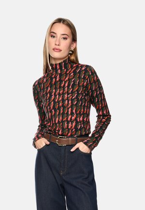 Woman wearing a dark turtleneck with red and pink abstract pattern, dark high-waist jeans, brown belt, and gold earrings, hands in pockets.
