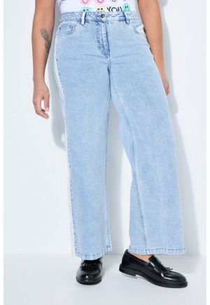 Hellblaue Jeans mit weitem Bein und weißen Seitenstreifen, getragen mit schwarzen Loafers und einem weißen T-Shirt mit Grafik, sichtbares Tattoo am linken Unterarm.