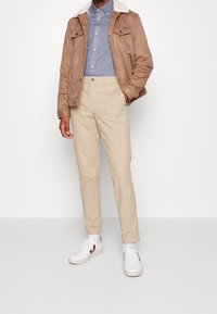 Chaqueta de gamuza marrón con cuello de pelo blanco, camisa celeste con botones, pantalones chinos beige y zapatillas blancas con detalles en rojo.