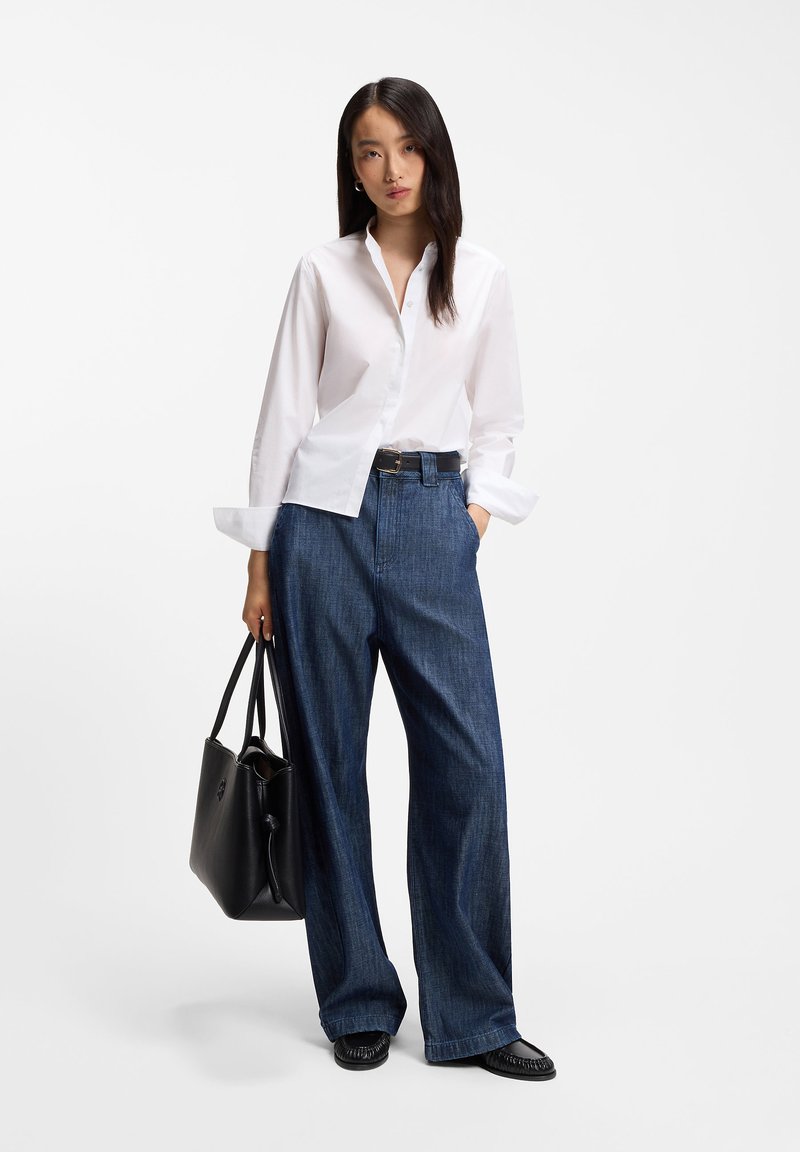 Femme portant une chemise blanche boutonnée, un jean bleu à jambes larges, une ceinture noire, des chaussures noires, et tenant un grand sac à main noir sur fond blanc.