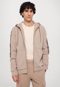 Sudadera beige con cremallera y un detalle texturizado en las mangas, con capucha ajustable y dos bolsillos frontales. Combinada con pantalones a juego.