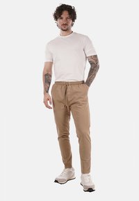 Uomo con capelli ricci e tatuaggi su entrambe le braccia, vestito con una t-shirt bianca, pantaloni beige con cordino e sneakers bianche, in piedi su sfondo bianco.