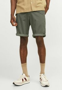 Shorts vert olive en tissu texturé, dotés de revers roulés. Associés à des baskets crème et des chaussettes beiges. Chemise à manches courtes visible.