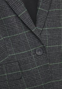 Blazer à carreaux gris foncé avec fines lignes vertes, col à revers pointu, bouton noir et poche à rabat à l'avant.