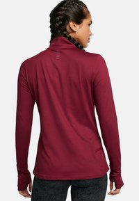 Maglia a maniche lunghe color mogano con colletto alto, texture liscia, fori per i pollici e una piccola tasca laterale; presenta un sottile dettagli di design sul retro.