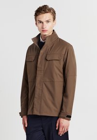 Giovane con capelli corti castano chiaro che indossa una giacca marrone con zip e tasche a pattina, in piedi contro uno sfondo chiaro e neutro.