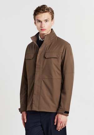 Junger Mann mit kurzem, hellbraunem Haar, der eine braune Jacke mit Reißverschluss und Klapptaschen trägt und vor einem schlichten, hellen Hintergrund steht.