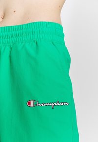 Zielone sportowe szorty z elastycznym pasem i bocznymi kieszeniami; posiadają czarne i czerwone haftowane logo "Champion". Gładka tekstura.