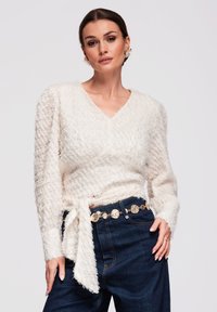 Femme portant un pull cache-cœur crème texturé avec des manches bouffantes, un jean en denim foncé et une ceinture à maillons dorés, posant devant un fond uni.