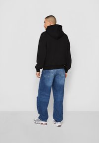 Sweatshirt noir à capuche avec une coupe décontractée, associé à un jean en denim oversized bleu clair et des chaussures de sport argentées.