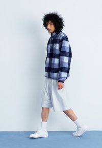 Pullover in pile di colore navy e blu chiaro a scacchi, con design a mezza zip. Indossato con pantaloni corti chiari e sneakers bianche.