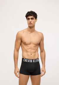 Juodos medvilnės bokso šortai su balta "CALVIN KLEIN" logotipo juosta. Modelis turi tonizuotą figūrą, demonstruodamas apatinio trikotažo pritaikymą ir dizainą.