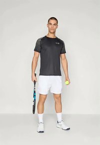 Musta urheilupaita, jossa on teksturoituneet harmaat olkapäät, yhdistettynä valkoisiin shortsit. Malli pitää kädessään tennismailaa ja keltaista tennispalloa.