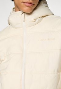 Doudoune Calvin Klein, beige clair, avec fermeture éclair sur le devant, capuche et panneaux cousus. Marque subtile près de la fermeture éclair de la même couleur.