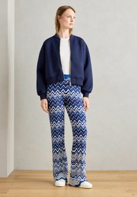 Navy cropped bomber jacket with a silver zipper, paired with blue and white zigzag-patterned flared pants and white sneakers.