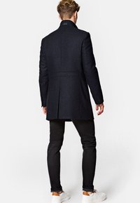 Czarny wełniany płaszcz o dopasowanym kroju, z wysokim kołnierzem i pojedynczym rozcięciem z tyłu, zestawiony z wąskimi czarnymi spodniami i białymi sneakersami.