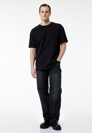 Młody mężczyzna stojący, ubrany w czarny luźny t-shirt, luźne ciemne jeansy i czarne buty, z jedną ręką w kieszeni na białym tle.
