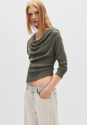 Maglione a coste verde oliva con scollatura a pieghe e maniche a tre quarti, abbinato a jeans azzurri a vita alta.