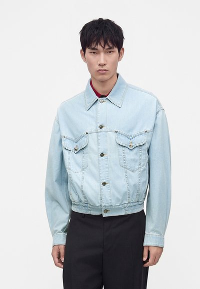 Jeune homme portant une veste en jean bleu clair avec des poches à boutons et un pantalon noir, debout devant un fond blanc.