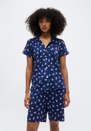 Moteris dėvi tamsiai mėlynos spalvos trumpomis rankovėmis marškinius su sagomis ir derančius šortus su mažais rožiniais ir baltais gėlių raštais, stovinti prieš baltą foną.