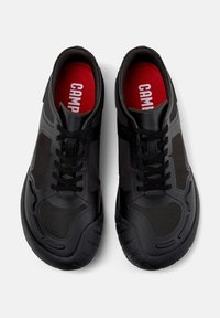Scarpe da ginnastica nere con un design in mesh testurizzato, punta arrotondata e un sistema di allacciatura evidente. L'interno presenta solette rosse con logo.
