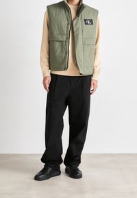 Zelená prošívaná vesta s nášivkou s logem, nošená přes béžovou mikinu. Kombinováno s černými kalhotami a černými teniskami. Hladká textura, neformální design.