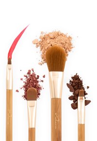 Set aus vier Makeup-Pinseln mit Bambusgriffen und goldenen Akzenten, begleitet von Häufchen loser Puder in Beigetönen, Rot und Braun.