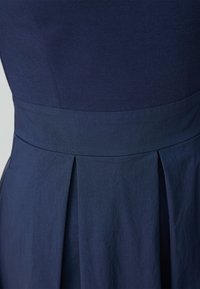 Marine jurkstof heeft een getailleerde lijfje met een gladde textuur en een geplooide rokdetail op de taille, wat de op maat gemaakte uitstraling benadrukt.